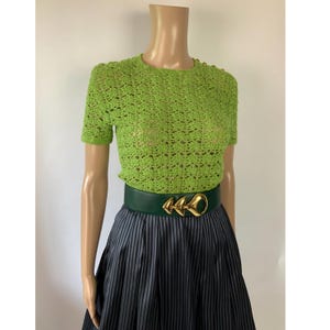 May include: A lime green short-sleeved crochet top with a round neckline, paired with a dark green leather belt featuring a gold-toned metal buckle with three triangular shapes, worn over a dark gray skirt with thin vertical pinstripes.