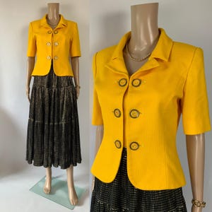 May include: A vintage two-piece suit. The bright yellow, short-sleeved blazer has four decorative buttons. The blazer is paired with a black and gold tiered skirt. The outfit is displayed on a mannequin.