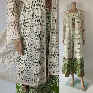 May include: A cream-colored, long-sleeved crochet cardigan with a floral pattern. The cardigan is worn over a green and pink floral print dress. The dress has a round neckline and long sleeves. The outfit is displayed on a mannequin.