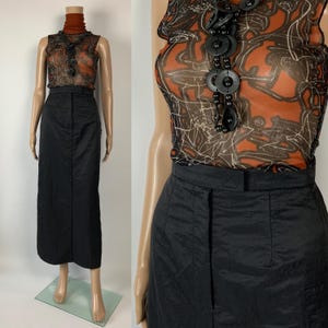 Puede incluir: Una falda lápiz negra combinada con una blusa transparente estampada y un cuello alto marrón. La blusa presenta un diseño naranja y negro, y un collar negro. El conjunto se exhibe en un maniquí.
