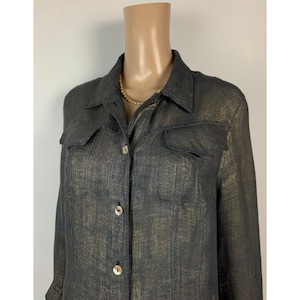 Può includere: Una camicia grigio scuro con bottoni e due tasche sul petto. La camicia ha una sottile lucentezza metallica dorata in tutto il tessuto. Il colletto e i polsini sono visibili e la camicia è abbottonata. La camicia è esposta su un manichino.