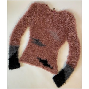 Könnte beinhalten: Ein staubrosa, flauschiger Langarmpullover mit schwarzen und grauen Akzenten an den Ärmeln. Der Pullover hat ein einzigartiges Design mit dunkelgrauen und schwarzen Formen auf der Vorderseite. Das Material wirkt weich und strukturiert.