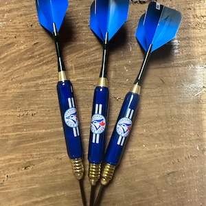 May include: Three blue darts with gold shafts and black tips. Each dart features a Toronto Blue Jays logo with the team's name and a bird graphic.