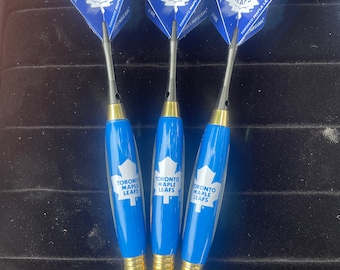 Pode incluir: Três dardos azuis e dourados com o logotipo dos Toronto Maple Leafs. Os dardos têm uma folha de bordo branca e as palavras "Toronto Maple Leafs" impressas neles.