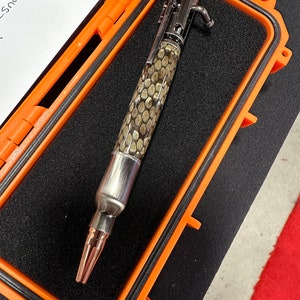 May include: A silver and brown pen with a snake skin pattern, in a black and orange case. The pen has a metal tip and a metal clip. The case has a black foam lining.
