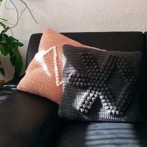 May include: Two crocheted throw pillows on a black couch. One pillow is a light pink color with a white beaded design. The other pillow is dark gray with a white textured design.