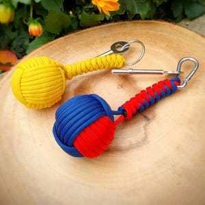 Puede incluir: Dos llaveros decorativos. Uno es amarillo, el otro es azul y rojo. Ambos tienen un diseño de bola anudada y están unidos a un llavero de metal o mosquetón. El fondo presenta flores naranjas y follaje verde.