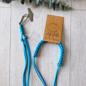 May include: A turquoise adjustable phone sling with a metal clip and a brown tag. The tag reads "Handmade by Daisy Dais" and "Adjustable Phone Sling". The sling is made of braided and knotted cord.