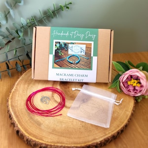 May include: A macrame charm bracelet kit featuring a brown box, pink cord, a silver charm, and a small white bag. The box is labelled "Handmade at Daisy Daisy" and "Macrame Charm Bracelet Kit."