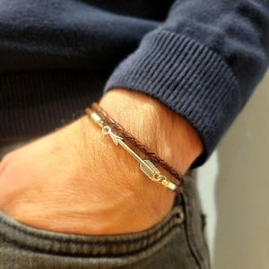 Puede incluir: Una pulsera de cuero marrón con un dije de flecha plateada. La pulsera es trenzada y tiene un cierre. El dije de flecha es detallado. La pulsera se lleva en la muñeca.