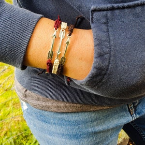 May include: Three braided bracelets with silver arrow charms. The bracelets are in burgundy, brown, and cream colours.