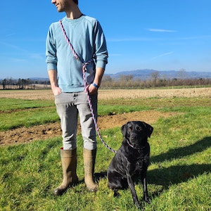 May include: A man wearing a light blue jumper and brown boots is standing in a field with a black Labrador Retriever. The dog is sitting and looking at the camera. The man is holding a blue, red and white rope lead.