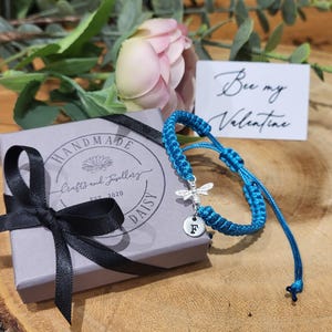 Puede incluir: Un brazalete de amistad azul turquesa con un dije de abeja de plata y un dije de inicial de plata. El brazalete está atado con un nudo y tiene una cola larga. El brazalete está empaquetado en una caja gris con una cinta negra y una etiqueta que dice "Handmade Crafts and Jewellery Daisy Est 2020". Una tarjeta con el texto "Bee my Valentine" está en el fondo.