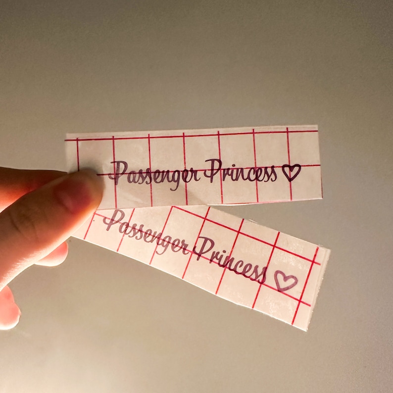 May include: Two white paper strips with a red grid pattern. The text "Passenger Princess" is written in cursive on each strip with a small heart symbol next to the text.