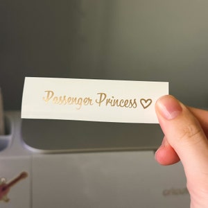 May include: A white card with the text "Passenger Princess" written in gold cursive lettering with a gold heart symbol.