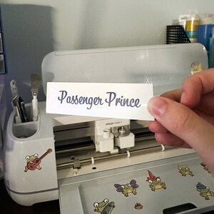 May include: A white sticker with the text "Passenger Prince" in blue cursive lettering. The sticker is being held in front of a Cricut Maker 3 machine.