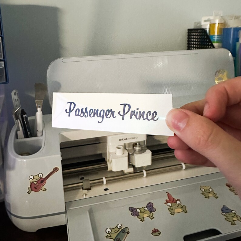 May include: A white sticker with the text "Passenger Prince" in blue cursive lettering. The sticker is being held in front of a Cricut Maker 3 machine.