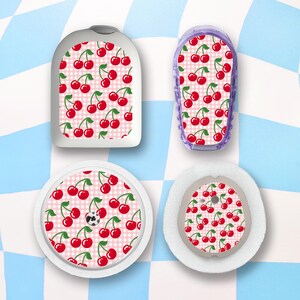 May include: Four medical device accessories with a cherry pattern on a pink gingham background. The accessories include a white rounded rectangle, a purple rectangular case, and two white circular devices. The cherries are red with green stems.
