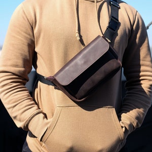 Puede incluir: Un bolso de cuero marrón cruzado con cierre de solapa y una correa de ante marrón. El bolso se lleva sobre una sudadera con capucha de color beige.