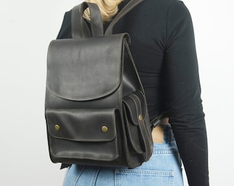 May include: A dark brown leather backpack with two front pockets and brass accents. The backpack is being worn by a person.