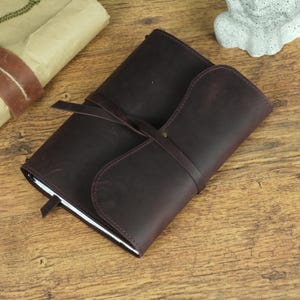 May include: A dark brown leather journal with a wrap-around cover design. The notebook features a long leather strap that secures the cover by wrapping around the exterior. The edges of the leather are smooth, and the surface shows a matte finish.