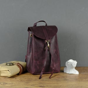 May include: A burgundy leather backpack with a flap closure and adjustable straps. The bag has a top handle and gold-colored hardware. A wrapped package and a small sculpture are also visible.