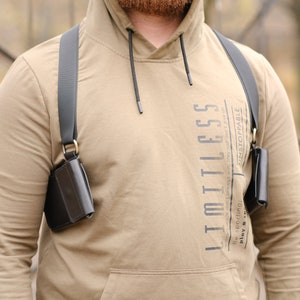 May include: A tan hooded sweatshirt with the text LIMITLESS printed vertically on the chest, worn with a black shoulder holster system featuring two dark leather pouches attached to adjustable black straps with metallic rings at the connection points.