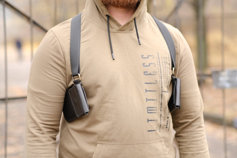May include: A tan hooded sweatshirt with the text LIMITLESS printed vertically on the chest, worn with a black shoulder holster system featuring two dark leather pouches attached to adjustable black straps with metallic rings at the connection points.