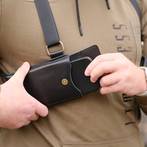 May include: A black leather chest holster featuring a vertical pouch with a brass snap button closure. The holster is attached to a black adjustable strap system that secures over the shoulders, designed to hold a smartphone securely against the chest.