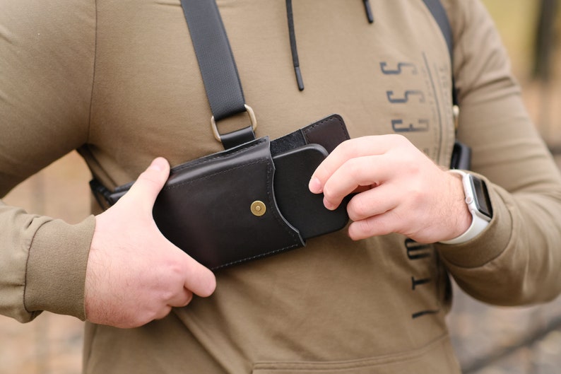 May include: A black leather chest holster featuring a vertical pouch with a brass snap button closure. The holster is attached to a black adjustable strap system that secures over the shoulders, designed to hold a smartphone securely against the chest.