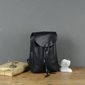 Op de afbeelding: Zwarte leren rugzak met een klepsluiting en trekkoord. Een verpakt pakket met een sleutel en een klein wit beeldhouwwerk staan naast de rugzak op een houten oppervlak. De achtergrond is effen grijs.