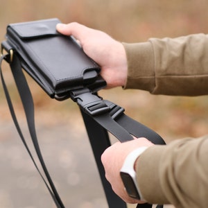 May include: A black leather phone holster with a flap closure and a matching black adjustable nylon strap. The holster features a plastic side-release buckle for secure attachment and a slim profile designed to hold a mobile device vertically.
