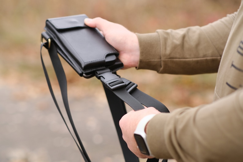 May include: A black leather phone holster with a flap closure and a matching black adjustable nylon strap. The holster features a plastic side-release buckle for secure attachment and a slim profile designed to hold a mobile device vertically.