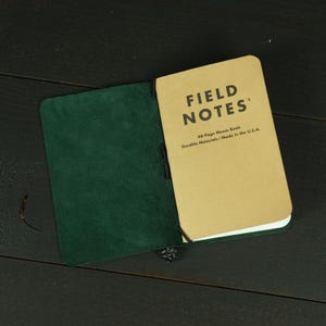 May include: A dark green leather notebook cover holding a tan Field Notes memo book. The cover features a smooth interior texture and a black elastic band securing the booklet. The tan booklet cover displays the text FIELD NOTES 48-Page Memo Book.