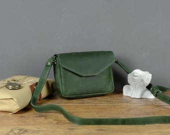 Pode incluir: Uma bolsa tiracolo verde-floresta com aba e alça de ombro ajustável. A bolsa apresenta um acabamento fosco liso e um formato retangular estruturado. Está posicionada ao lado de um pacote embrulhado e uma pequena escultura de pedra branca.