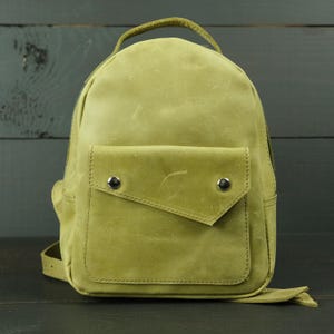 Könnte beinhalten: Ein hellolivgrüner Lederrucksack mit glatter, matter Oberfläche, einem Tragegriff oben und einer Vordertasche mit asymmetrischer Klappe, die mit zwei silberfarbenen Metalldruckknöpfen gesichert ist. Das Design weist sichtbare Nähte an den Kanten auf.