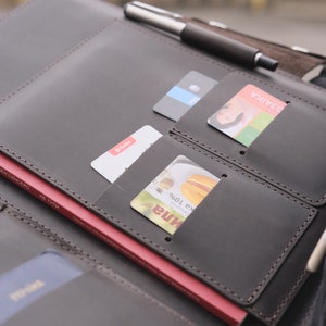 Puede incluir: Una carpeta organizadora de cuero marr&oacute;n oscuro con m&uacute;ltiples ranuras para tarjetas, un portal&aacute;pices con un bol&iacute;grafo plateado y negro, y un bolsillo grande que contiene un libro rojo. La carpeta est&aacute; cosida con hilo oscuro y contiene tarjetas.