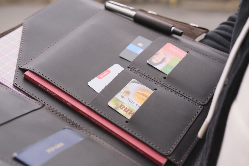Puede incluir: Una carpeta organizadora de cuero marr&oacute;n oscuro con m&uacute;ltiples ranuras para tarjetas, un portal&aacute;pices con un bol&iacute;grafo plateado y negro, y un bolsillo grande que contiene un libro rojo. La carpeta est&aacute; cosida con hilo oscuro y contiene tarjetas.