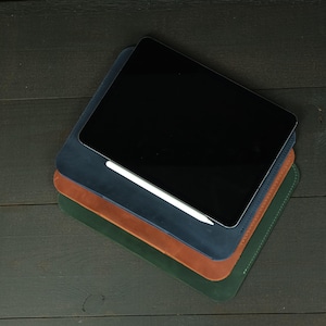 May include: A stack of three rectangular leather sleeves in navy blue, tan, and dark green. A black tablet with a glossy screen rests on the top navy blue sleeve, with a white stylus pen positioned horizontally across the surface of the leather.