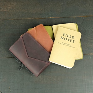 May include: A dark purple leather notebook cover with a V-shaped flap and black elastic cord closure, layered over a tan leather cover and a beige Field Notes memo book with black text reading Field Notes 48-Page Memo Book.