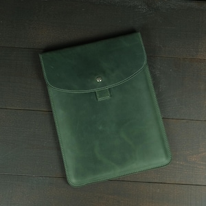 May include: A dark forest green leather sleeve with a smooth matte finish, featuring a rounded flap closure secured by a single silver-toned metal snap button. The rectangular case has stitched edges and a clean, minimalist construction.