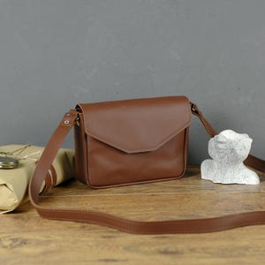 May include: A brown leather crossbody bag with a flap closure and adjustable strap. The bag is displayed on a wooden surface with a wrapped package and a small statue.