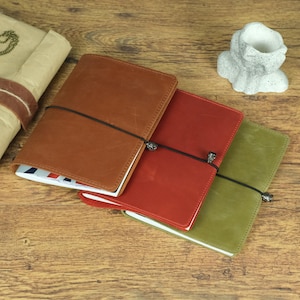 Puede incluir: Tres cuadernos rectangulares con cubierta de cuero en color tostado, rojo y verde oliva. Cada cuaderno está asegurado con un cordón elástico negro que presenta una pequeña cuenta metálica en forma de calavera. Están apilados ligeramente.