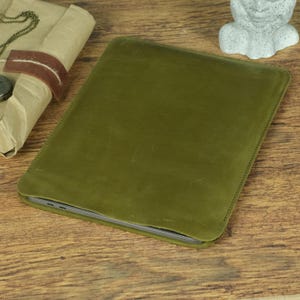Pode incluir: Uma capa retangular de couro verde-oliva projetada para um tablet ou laptop. A capa apresenta costuras visíveis ao longo das bordas, um acabamento fosco suave com sutis variações naturais na superfície e um perfil fino e protetor.
