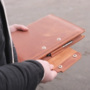 Puede incluir: Una carpeta de cuero color canela con una solapa plegable y dos cierres de presi&oacute;n de metal plateado. El exterior incluye una trabilla de cuero para sujetar un bol&iacute;grafo plateado, colocado horizontalmente a lo largo del borde frontal.