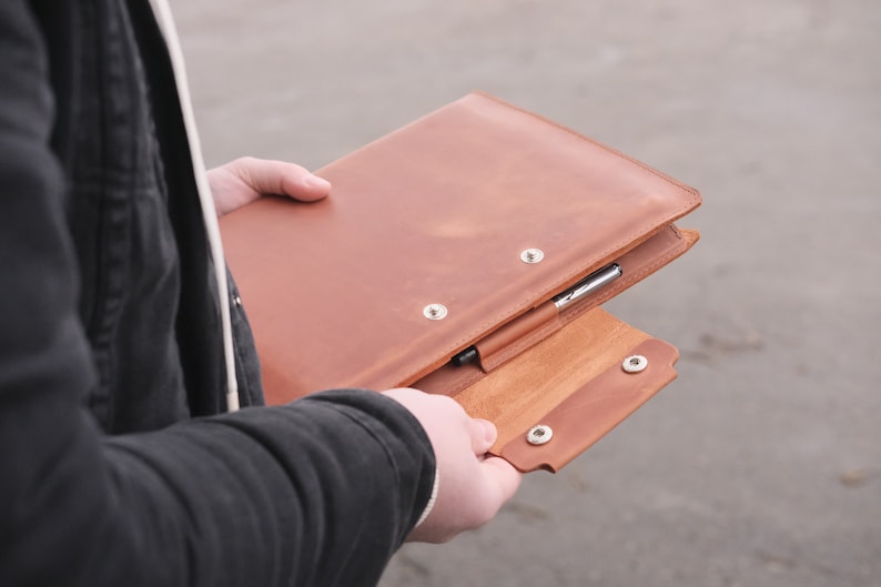 Puede incluir: Una carpeta de cuero color canela con una solapa plegable y dos cierres de presi&oacute;n de metal plateado. El exterior incluye una trabilla de cuero para sujetar un bol&iacute;grafo plateado, colocado horizontalmente a lo largo del borde frontal.