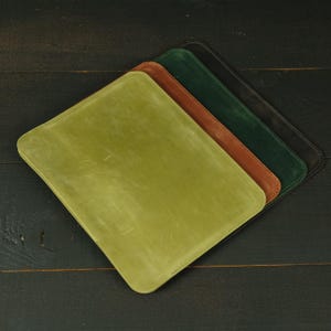 Pode incluir: Uma pilha de quatro capas retangulares de couro com cantos arredondados, dispostas num padrão em cascata. As capas são de cor verde-oliva claro, castanho médio, verde-floresta escuro e preto, cada uma com um acabamento mate e liso.