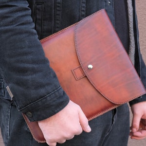 Peut inclure: Une pochette à documents en cuir marron dotée d'un rabat arrondi avec un bouton-pression en métal argenté. La pochette présente des coutures visibles sur les bords et une patte en cuir carrée centrée sur la fermeture du rabat avant.