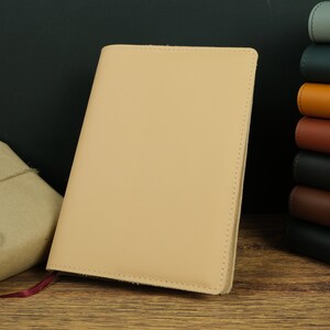 May include: A light beige leather notebook with a red ribbon bookmark. The notebook is closed and sitting on a wooden surface.