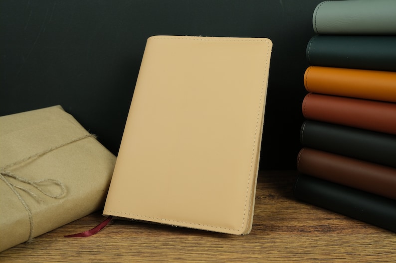 May include: A light beige leather notebook with a red ribbon bookmark. The notebook is closed and sitting on a wooden surface.